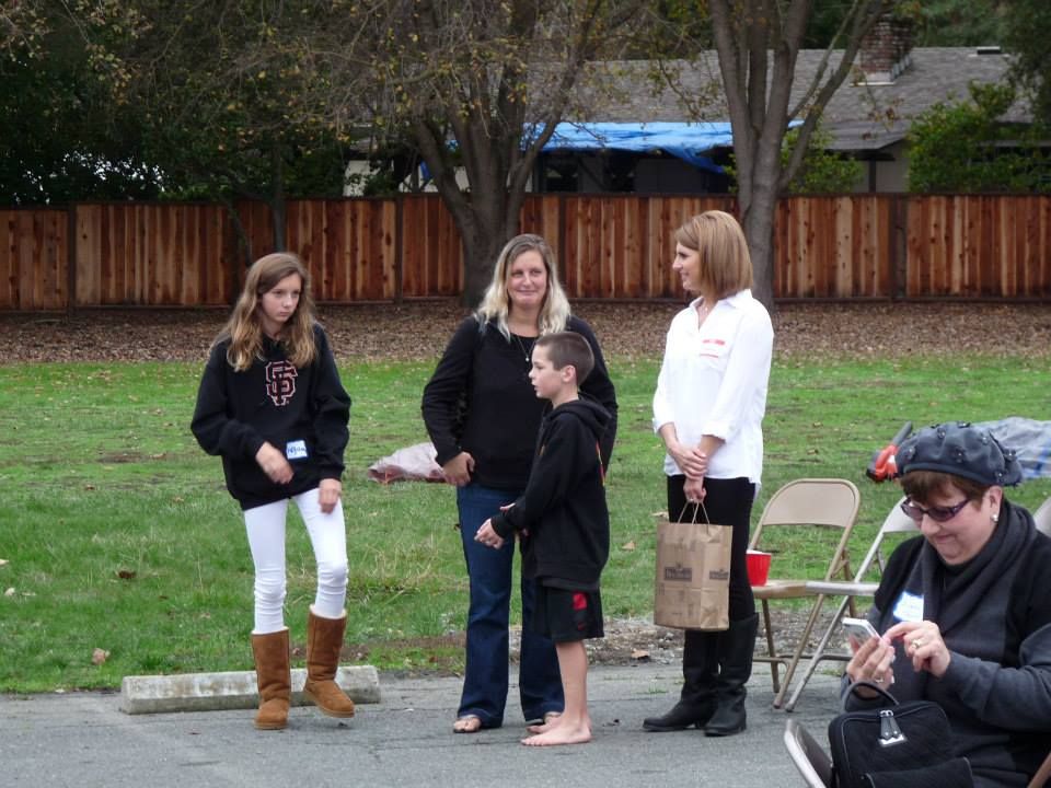 A group of people standing outdoors on an asphalt lot near a grass lawn and a wooden fence, with one person using a phone.