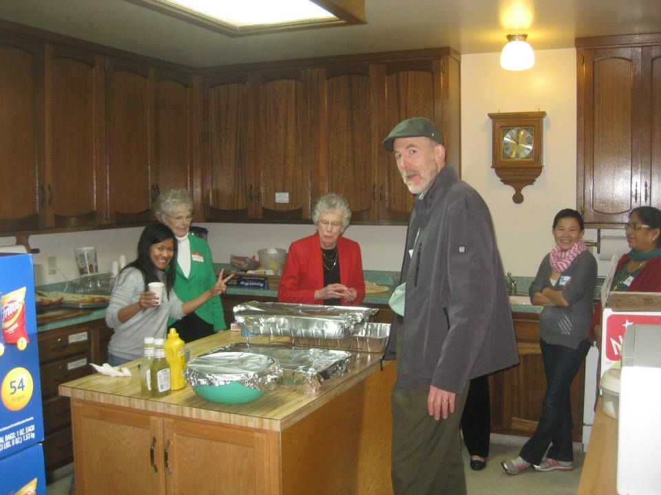 People gathered in a kitchen around a food-covered island; some are smiling and gesturing while preparing for a meal.