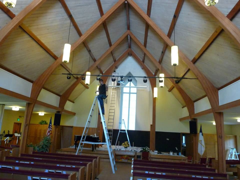 A person on a ladder adjusts lights hanging from the vaulted wooden ceiling of a church sanctuary.