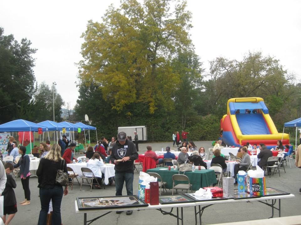 An outdoor community event features tables with blue canopies, a large inflatable slide, and people socializing.