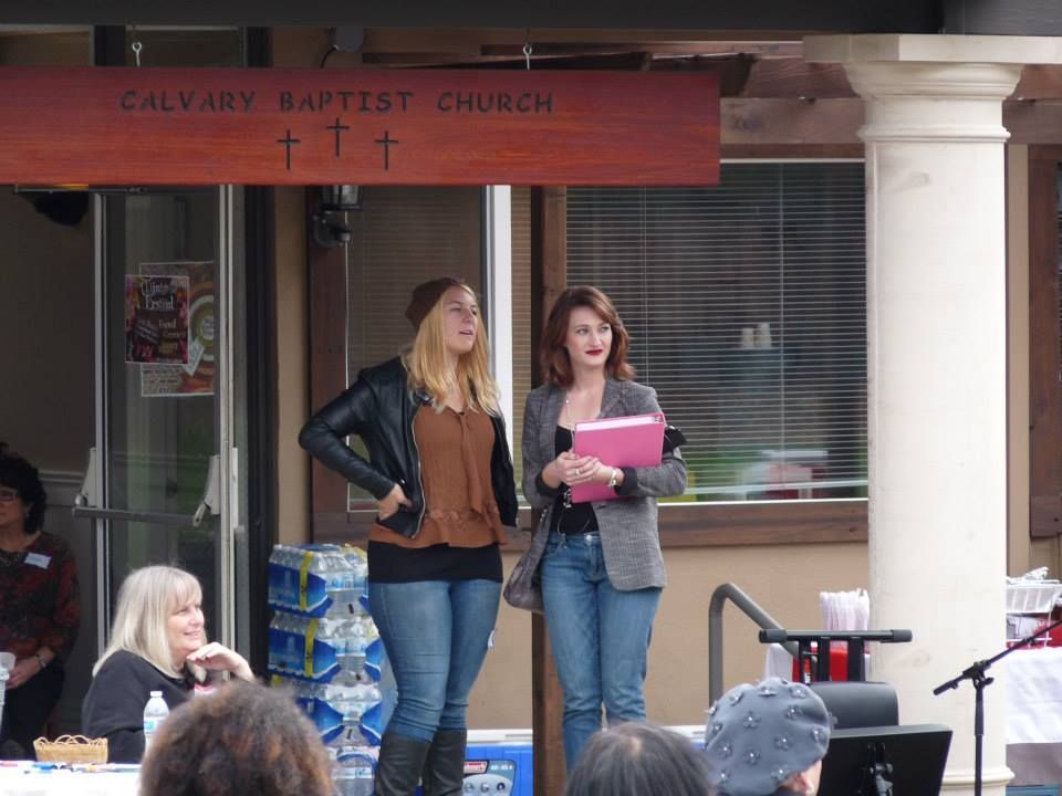 Two people stand in front of Calvary Baptist Church; one holds a pink folder, while another person sits at a nearby table.