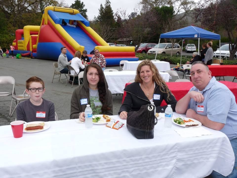Four people sit at a white outdoor table with food, with an inflatable slide and parking lot in the background.