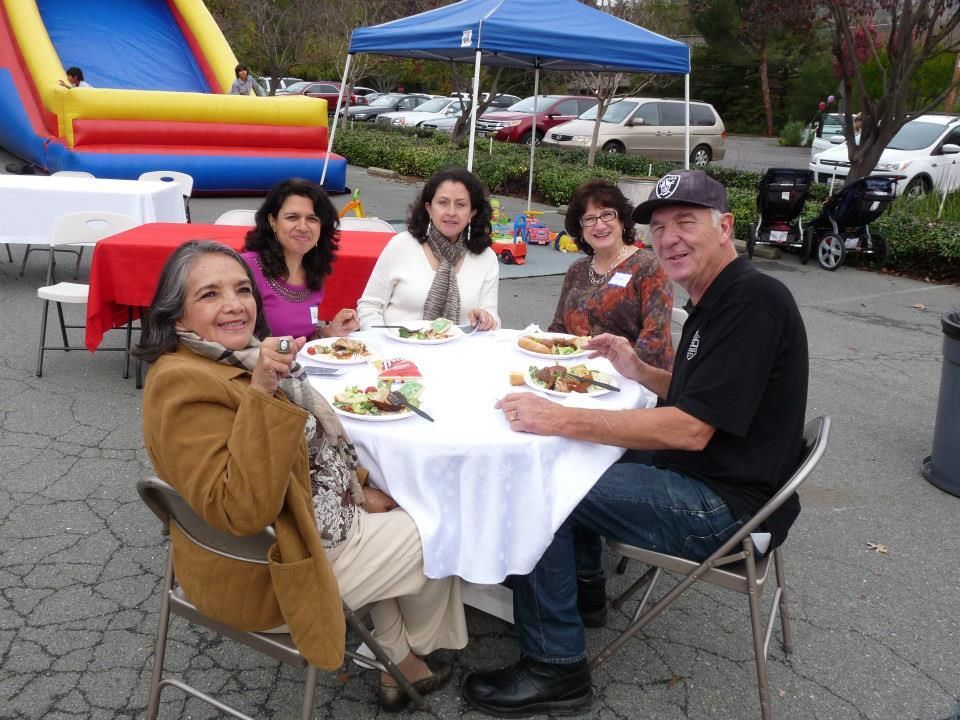 Four people sitting around a round table outdoors, smiling while eating at a community gathering.