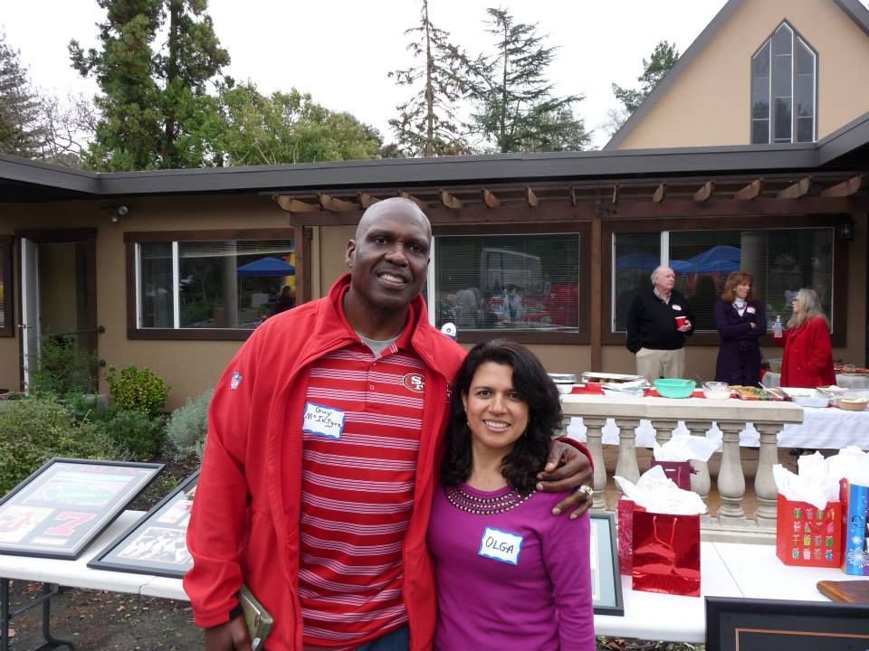 Two people pose for a photo at an outdoor gathering near a table with gift bags and refreshments in front of a building.