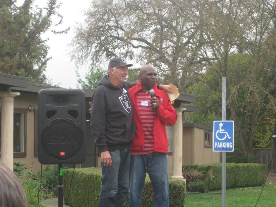 Two people stand outdoors near a speaker and a handicap parking sign; one wears a Raiders hoodie, the other a red jacket.