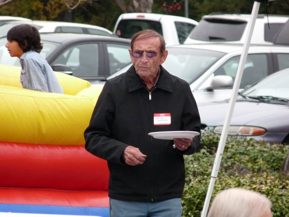 An individual wearing a black jacket and sunglasses holds a white plate at an outdoor event near a bounce house.