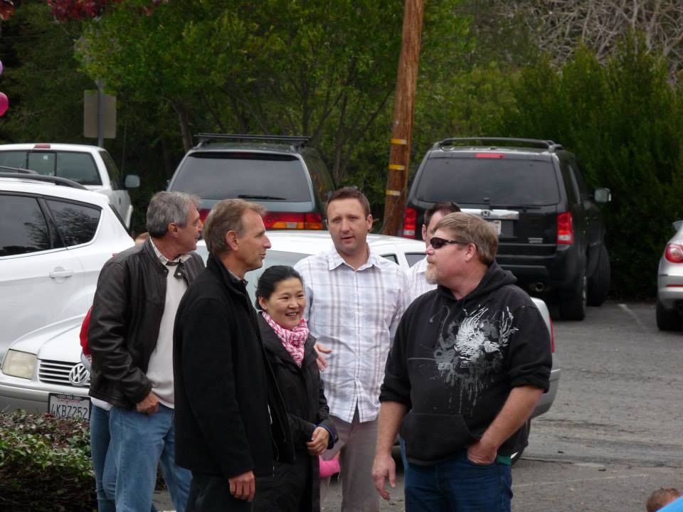 A group of people talking in a parking lot filled with various cars on a cloudy day.