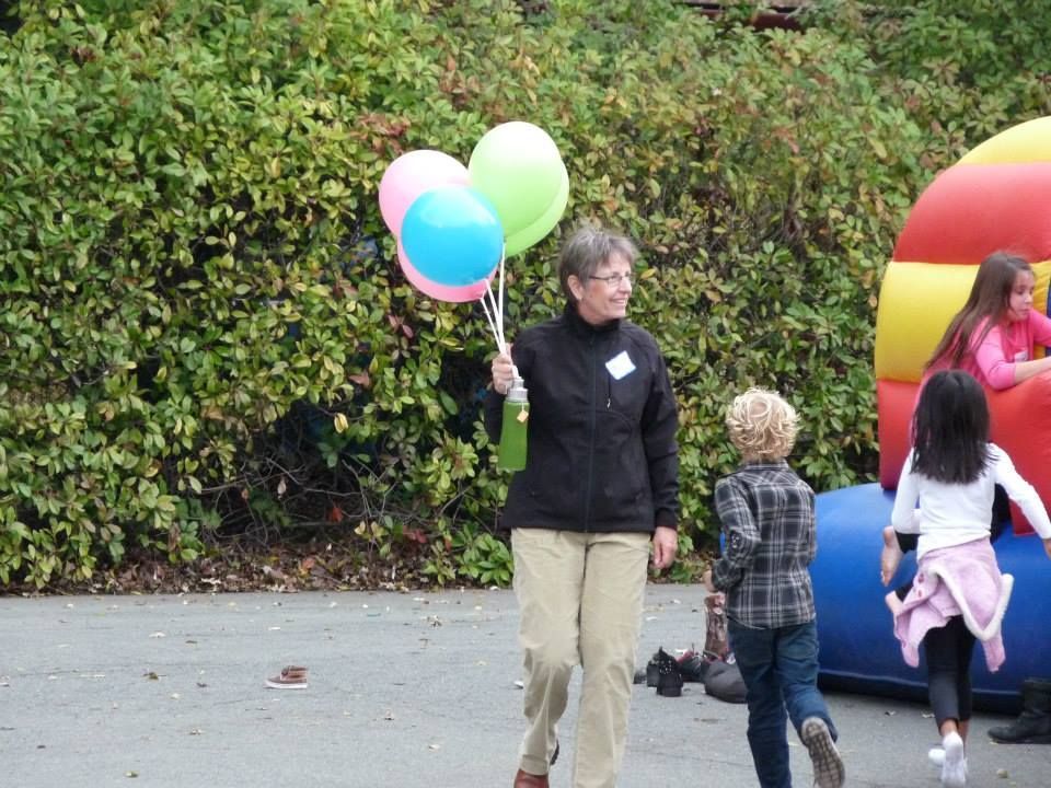 A person carries three balloons near a bouncy castle where children play outdoors in front of a hedge.