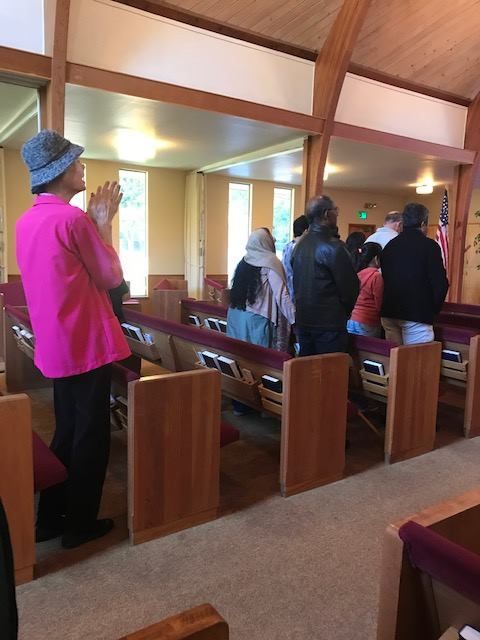 People stand in wooden pews inside a church, some with hands clasped in prayer during a service.