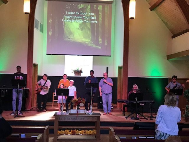 A musical worship band performs on a church stage under green lights with lyrics projected on a screen above them.
