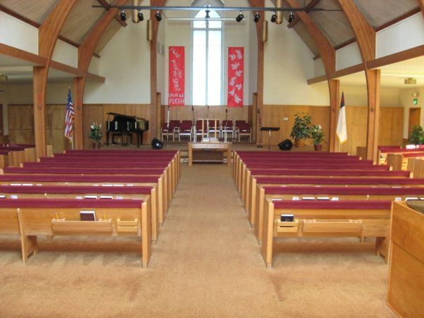 A view down the center aisle of a church sanctuary with rows of wooden pews, a carpeted floor, and a stage in the back.