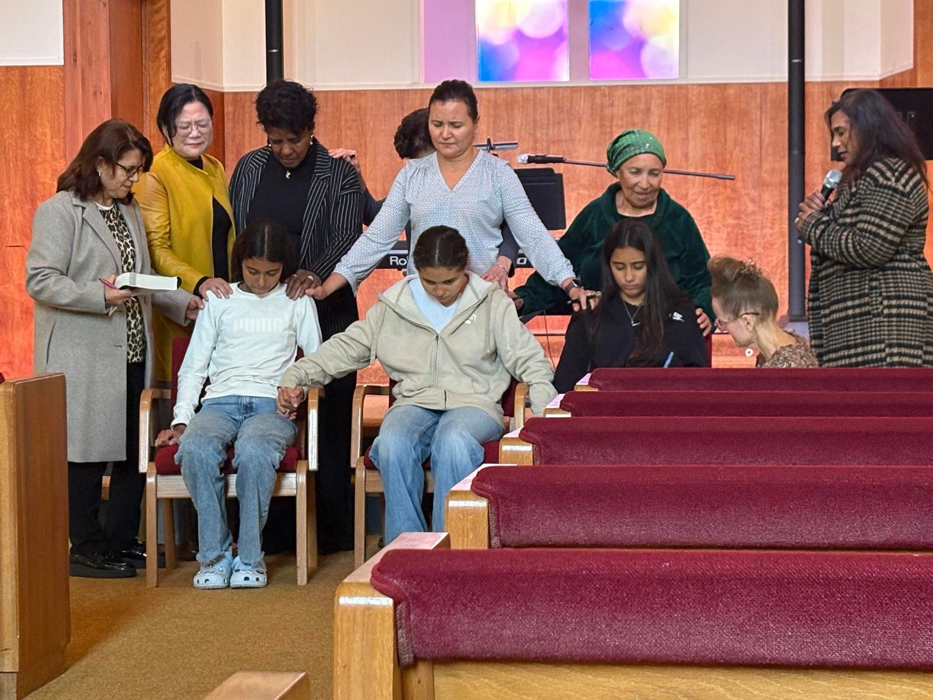 A group stands in a church interior, some placing their hands on two seated people to offer a prayer or blessing.