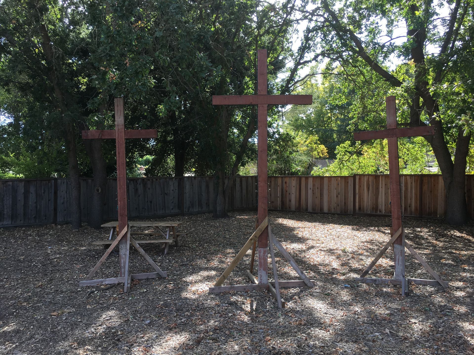 Three wooden crosses stand in a line on a wood-chipped surface in front of a wooden fence and leafy trees.