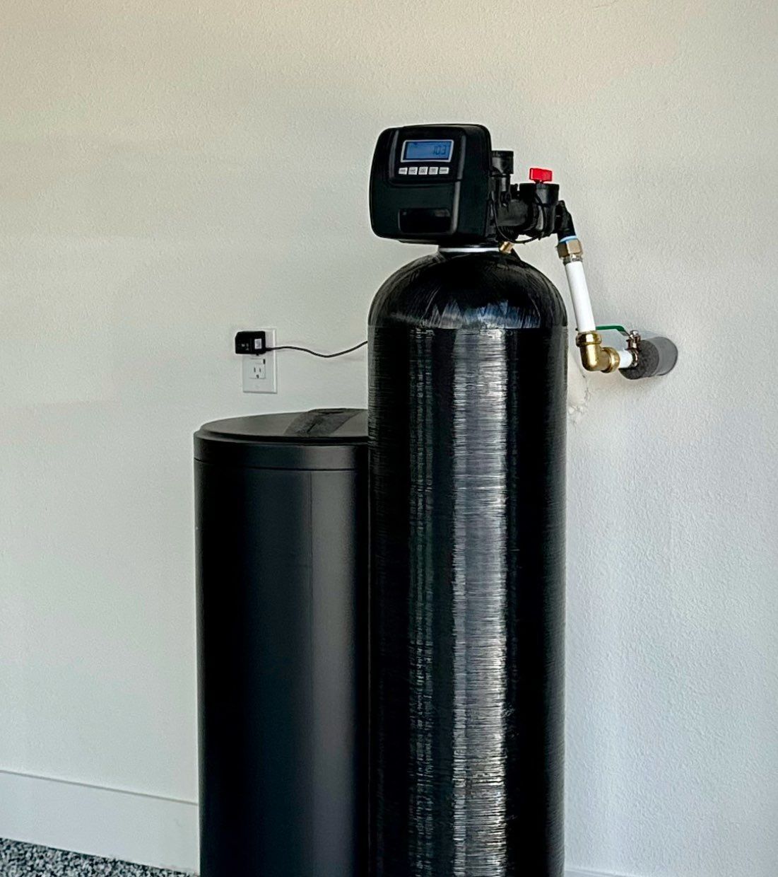 A black water softener is sitting next to a white wall.