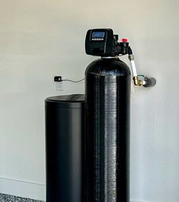 A black water softener is sitting next to a white wall.