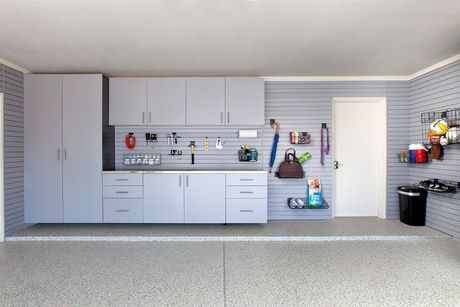 Light grey garage cabinets and storage shelving installed on slatted wall panels with a white door and epoxy floor.