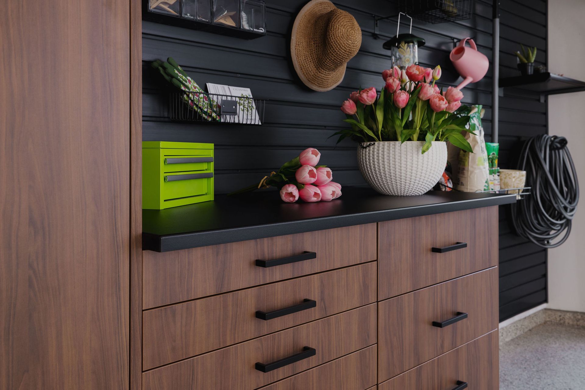 A wooden cabinet with black drawers sits against a black slat wall featuring shelves, a straw hat, and a potted tulip.