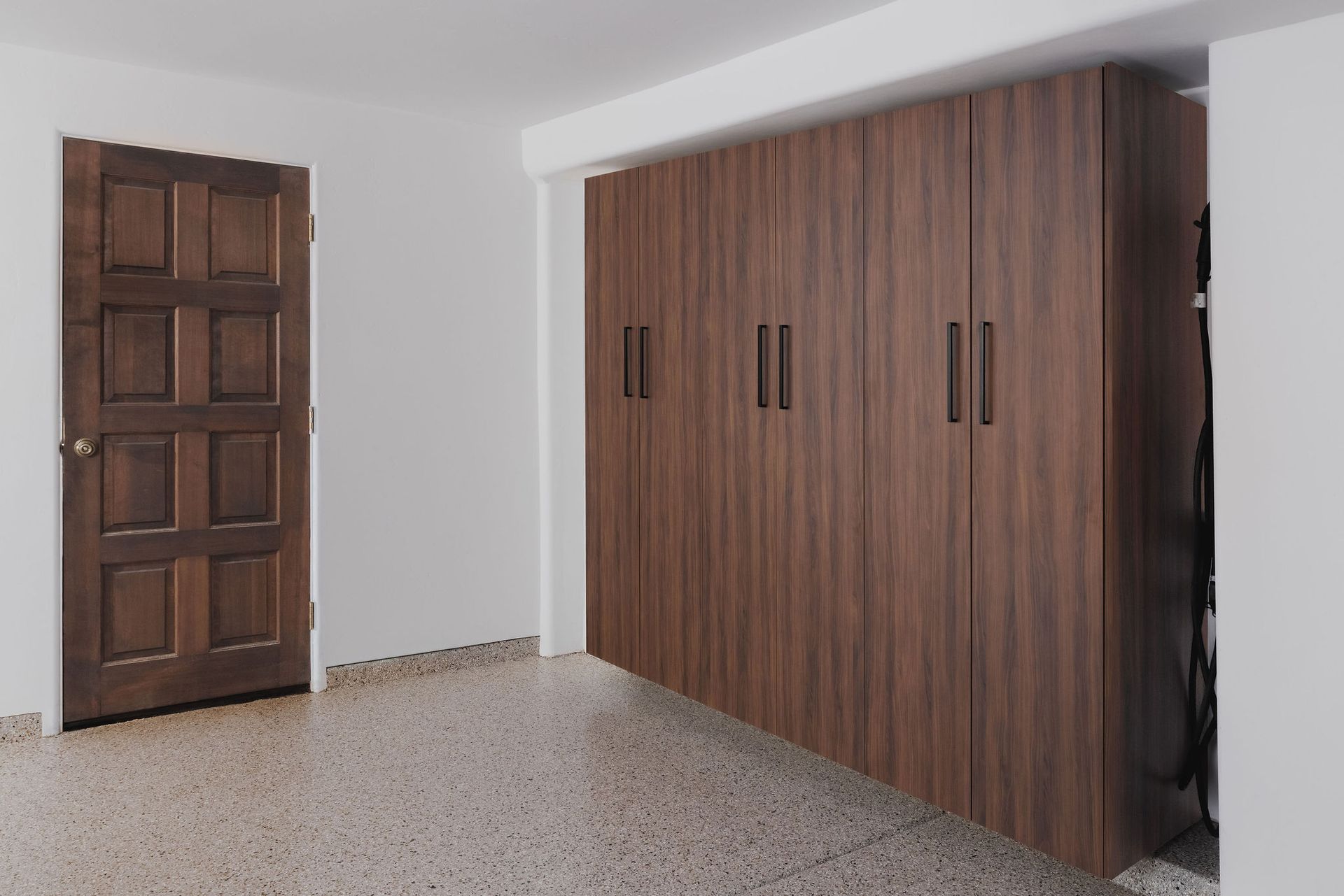 A dark wood-paneled door stands beside a matching, wall-to-wall storage unit in a room with speckled flooring.