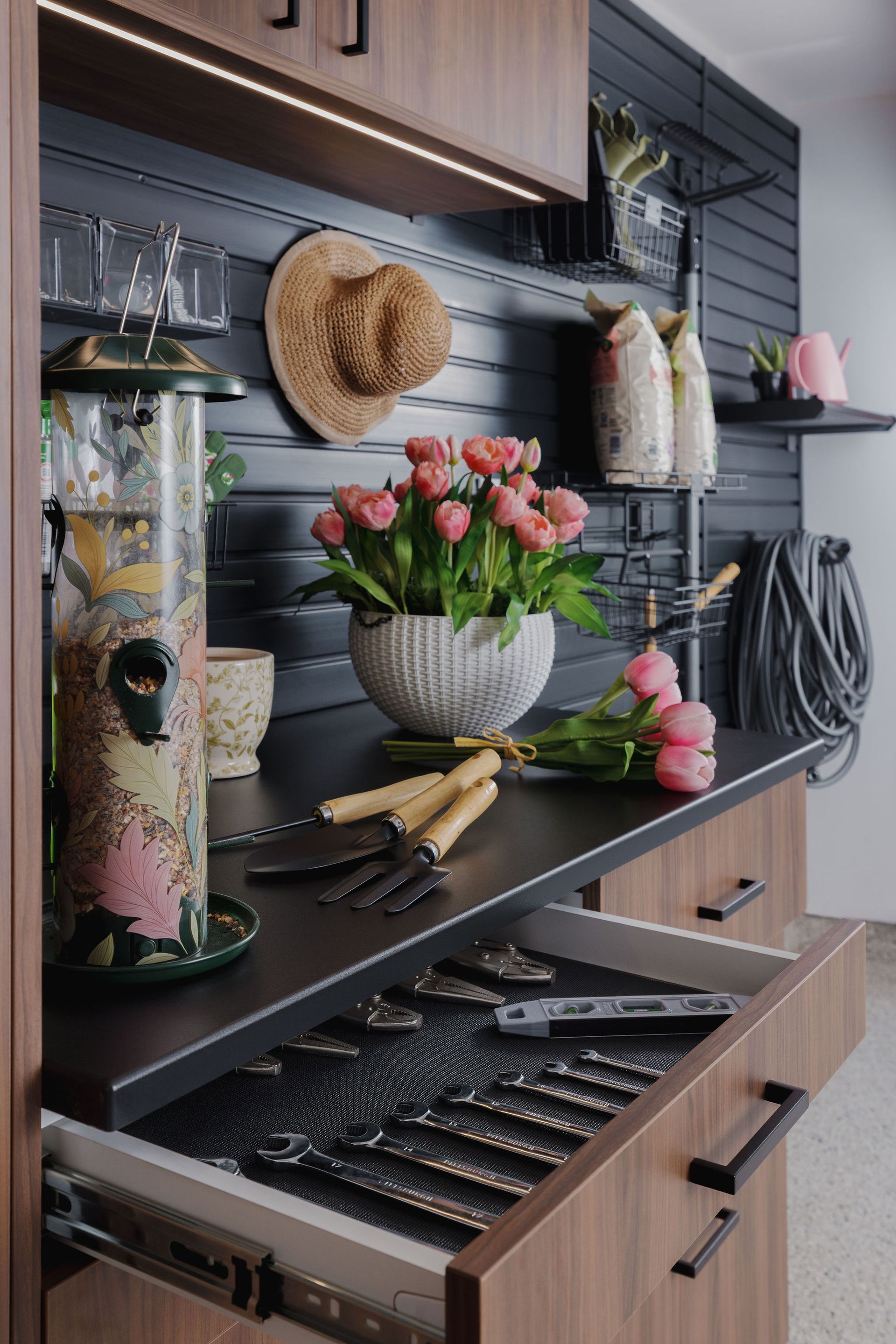 A potting bench with an open drawer of gardening tools, a bird feeder, a bowl of pink tulips, and wall-mounted storage.