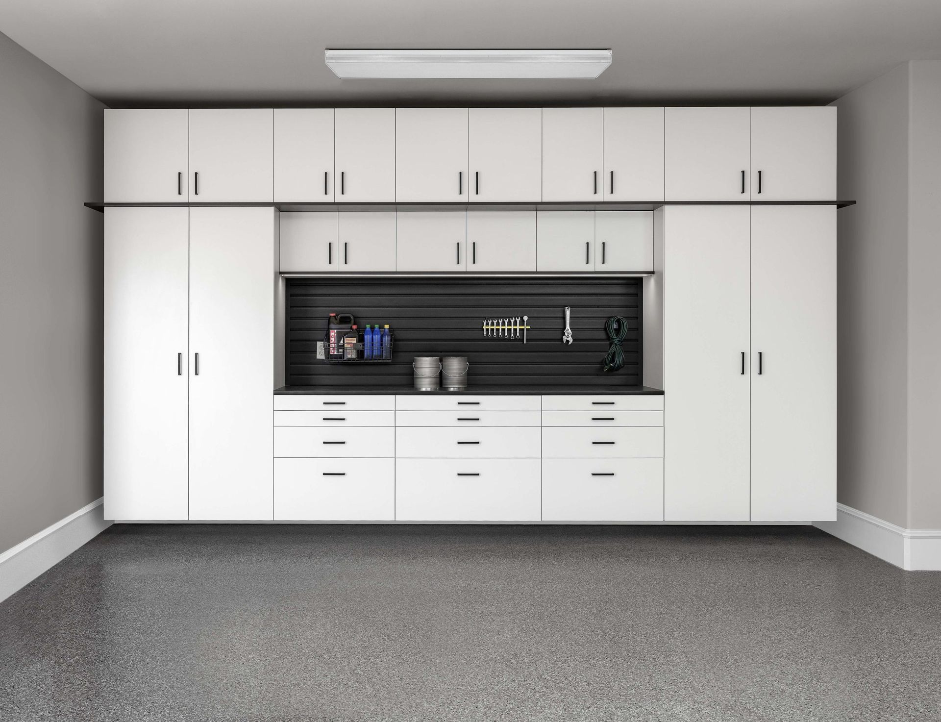 White garage storage cabinets with drawers and an open middle workspace against a gray wall and speckled concrete floor.