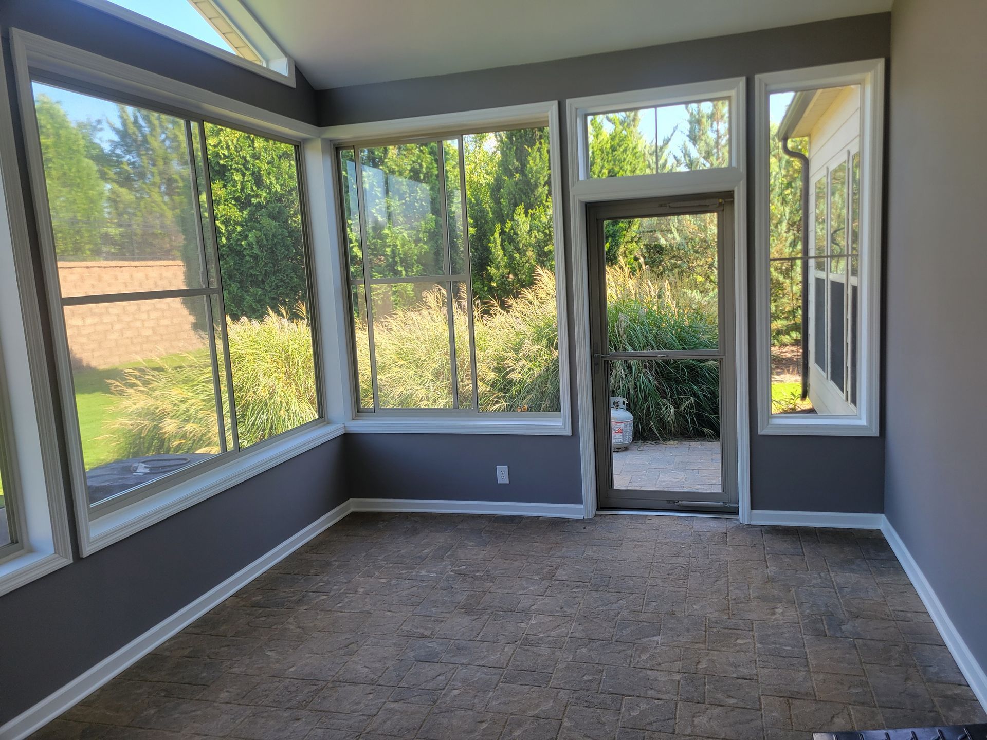 Empty sunroom with gray walls, large windows, and a glass door opening to a garden.