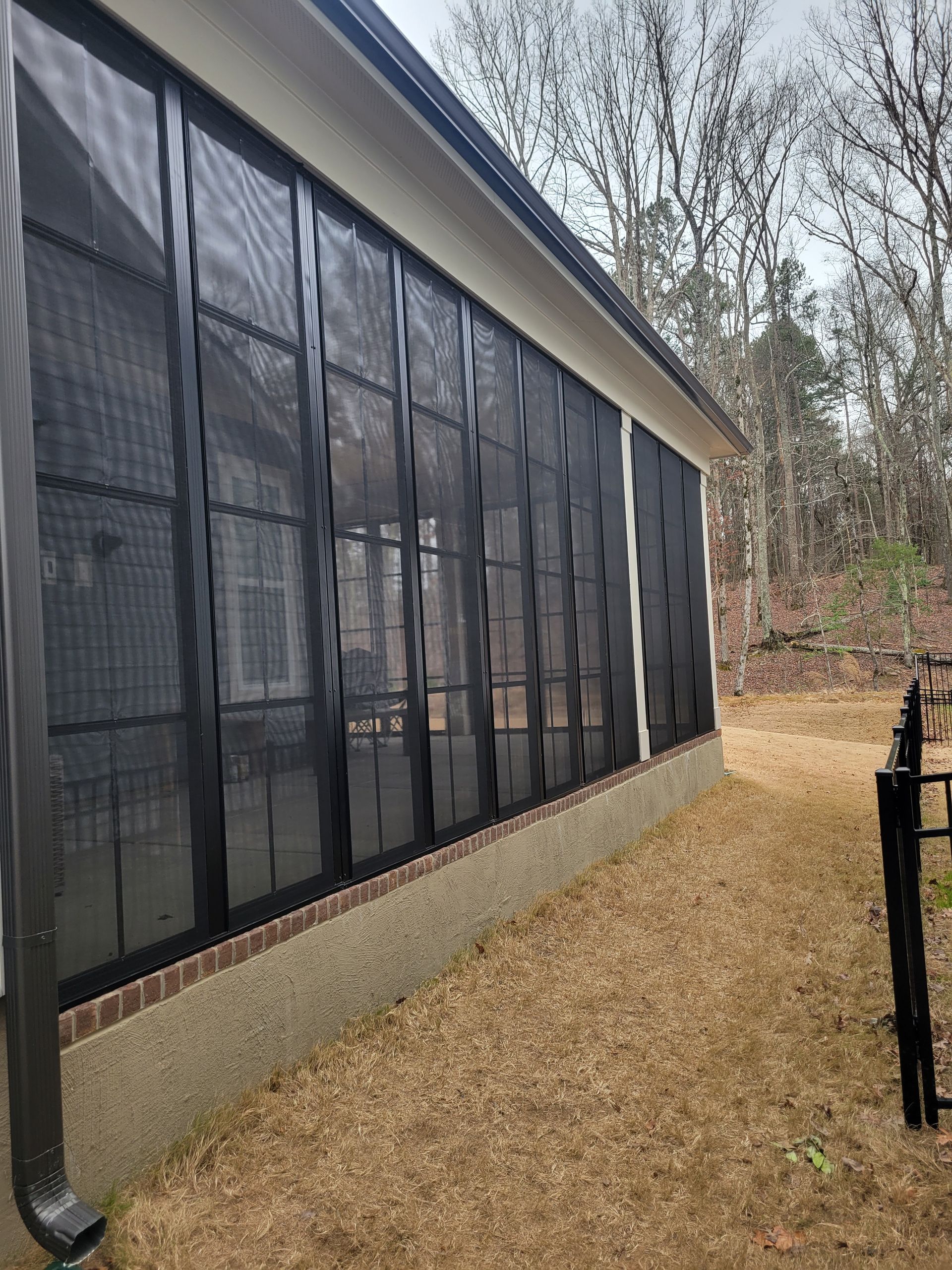 Side of a house with black screened porch panels and tan gravel beside leafless trees.