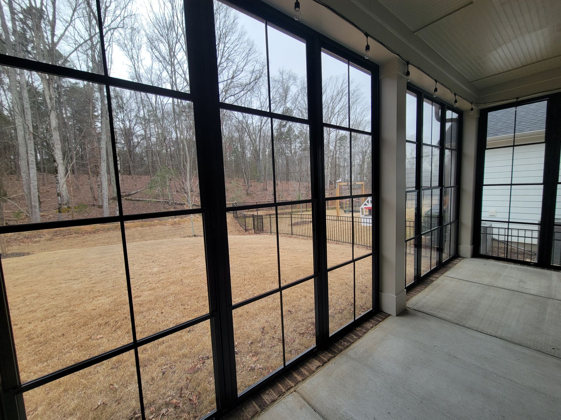 Screened porch with black-framed windows overlooking a bare, wooded yard and tan ground