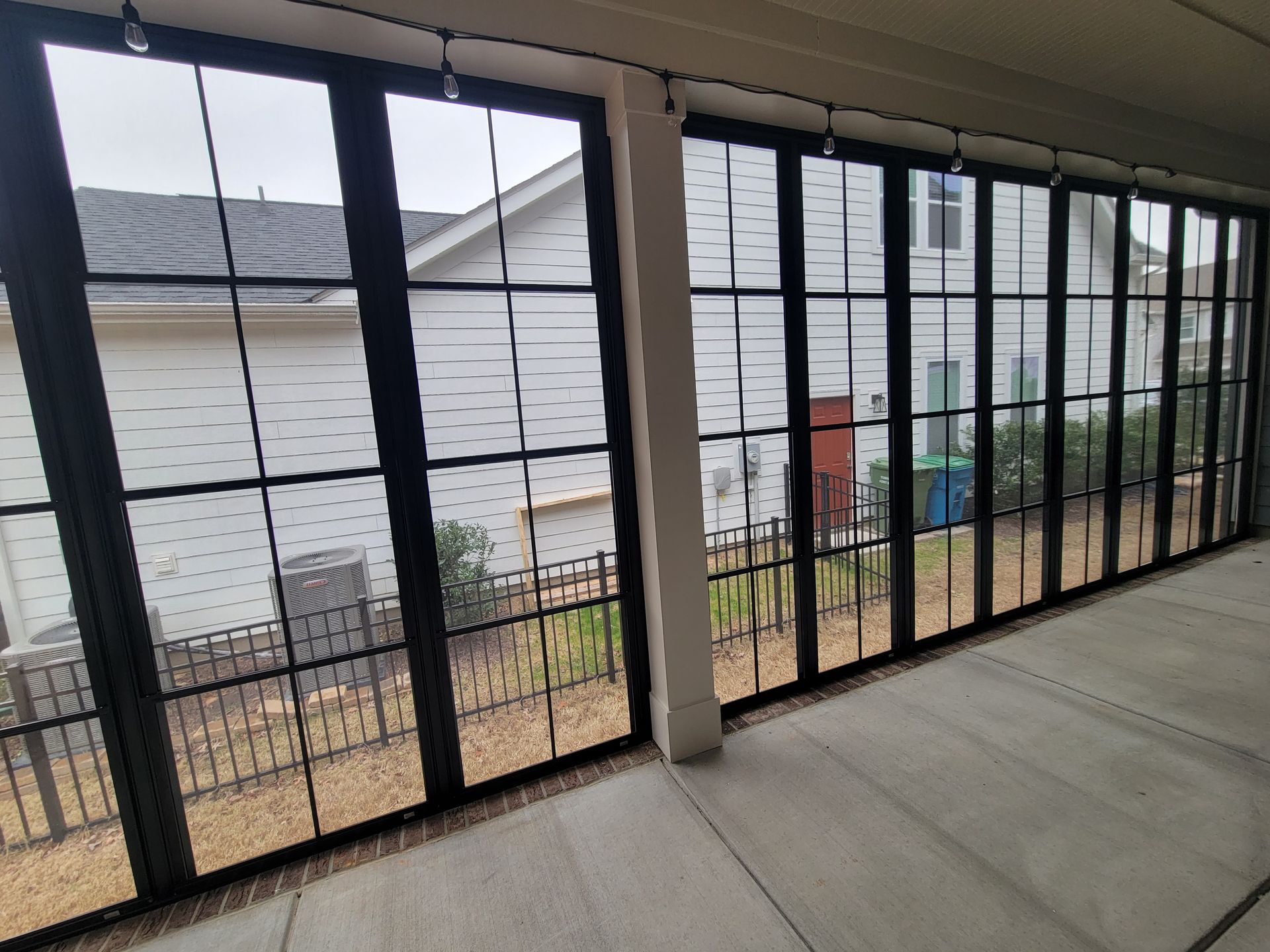 Empty screened porch with black metal railing, overlooking a white house and yard through large windows.