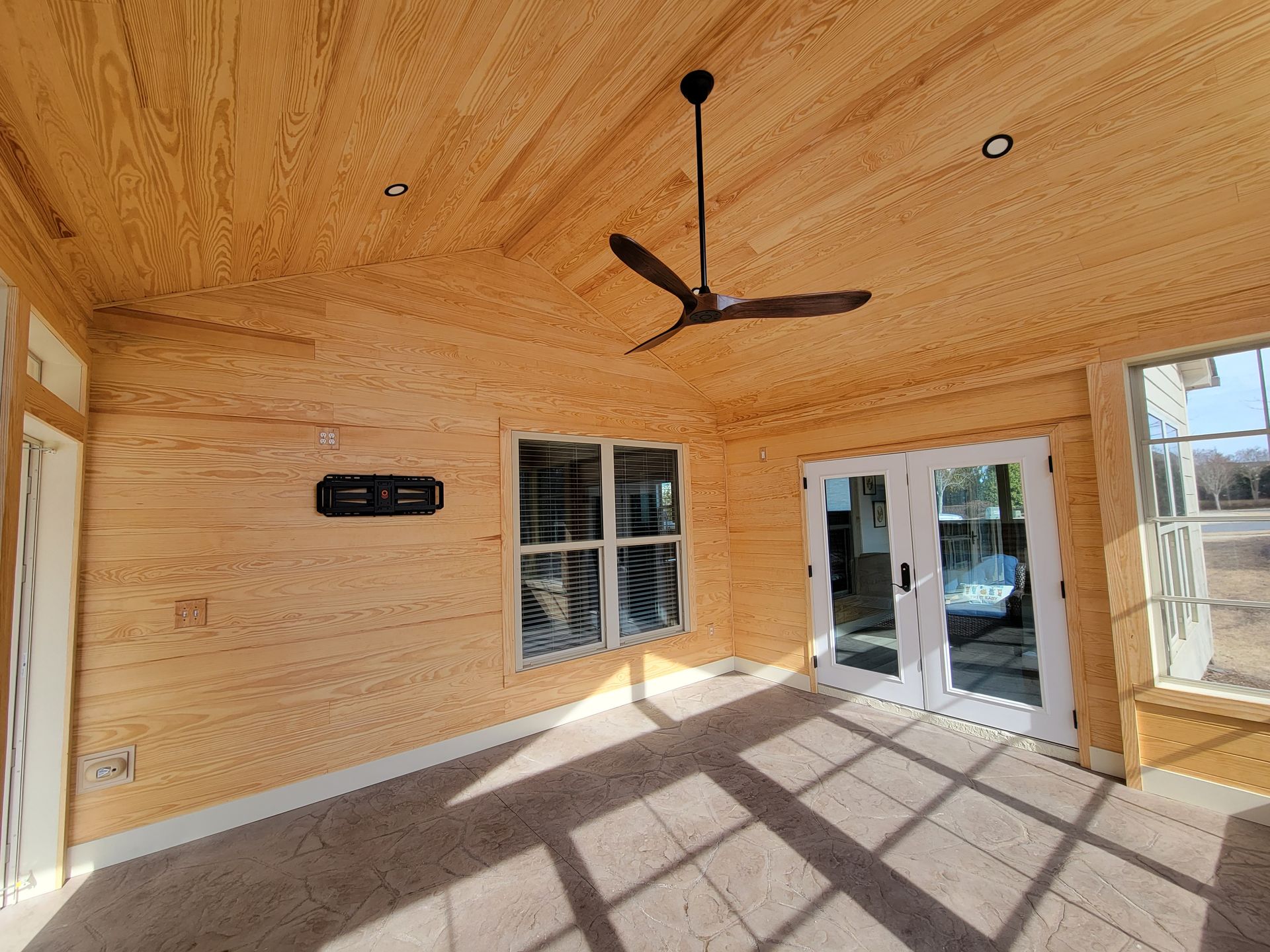 Empty screened-in porch with wood-paneled walls, ceiling fan, and sliding glass doors casting shadows on the floor