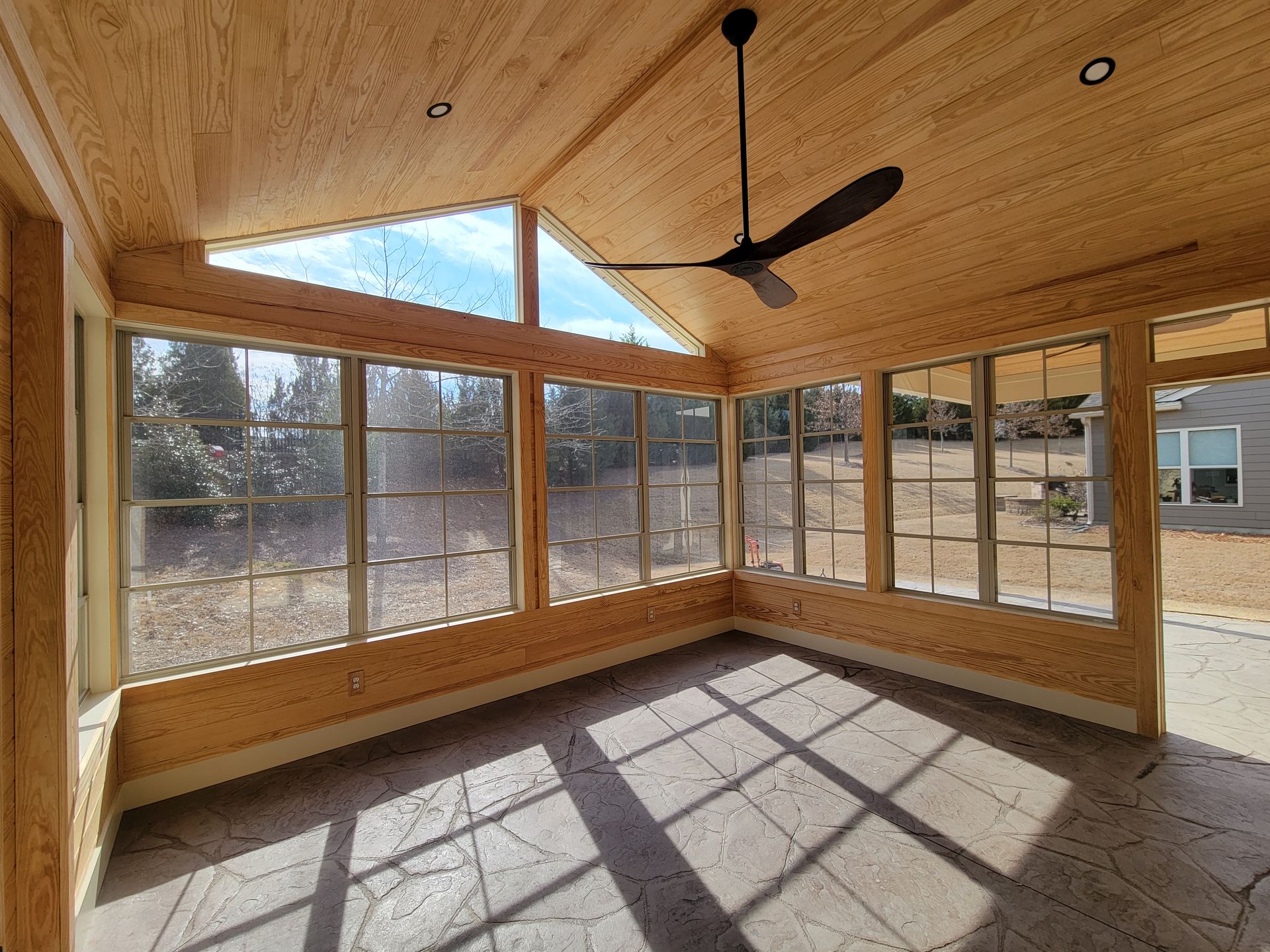 Sunlit enclosed porch with wood-paneled ceiling, large windows, and a ceiling fan casting grid shadows on the floor