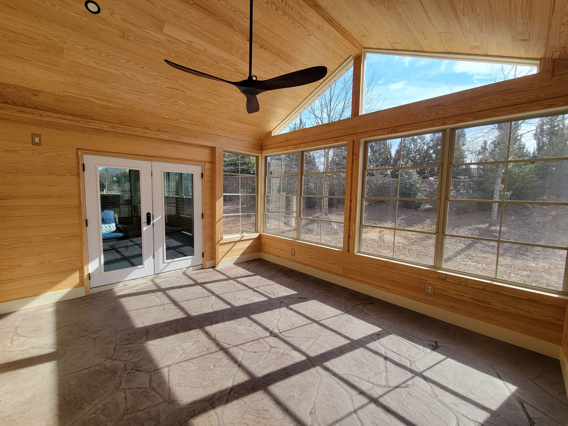 Bright sunroom with wood walls, large windows, ceiling fan, and tiled floor casting long shadows.