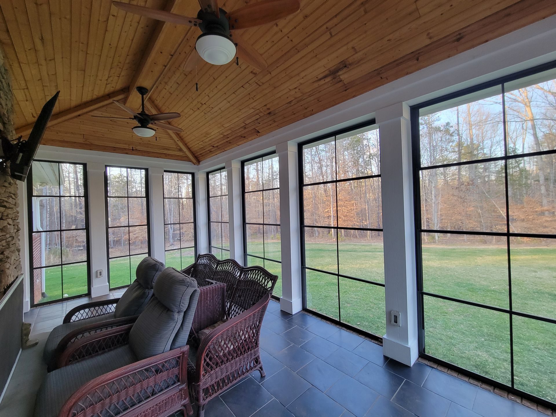 Sunroom with wicker chairs, wood ceiling, and large screened windows overlooking a grassy yard.