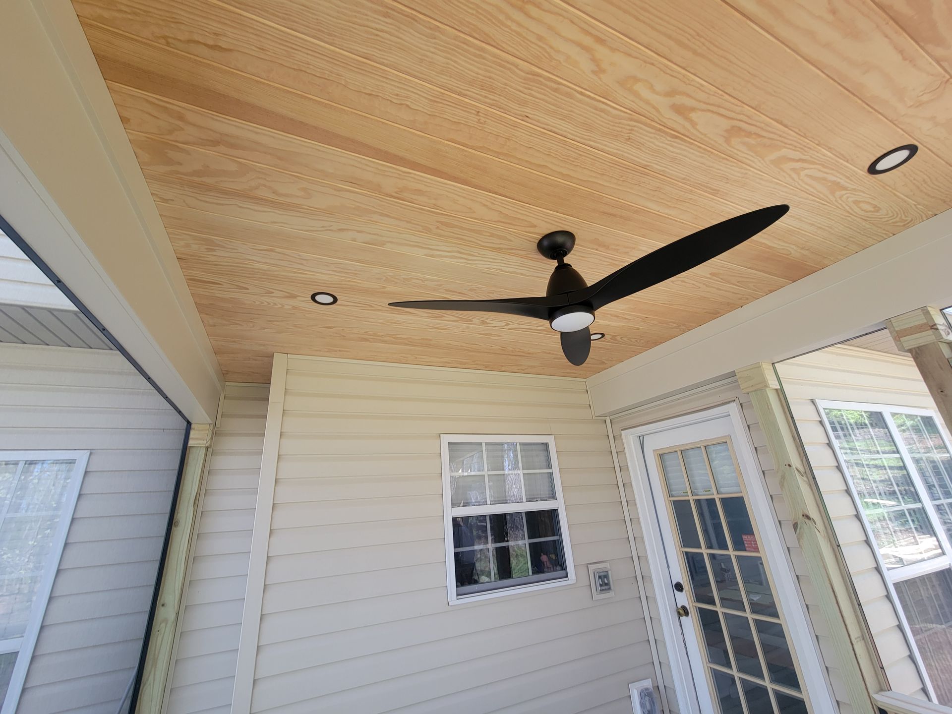 Covered porch with wood ceiling, black ceiling fan, recessed lights, and white siding with windows and glass door
