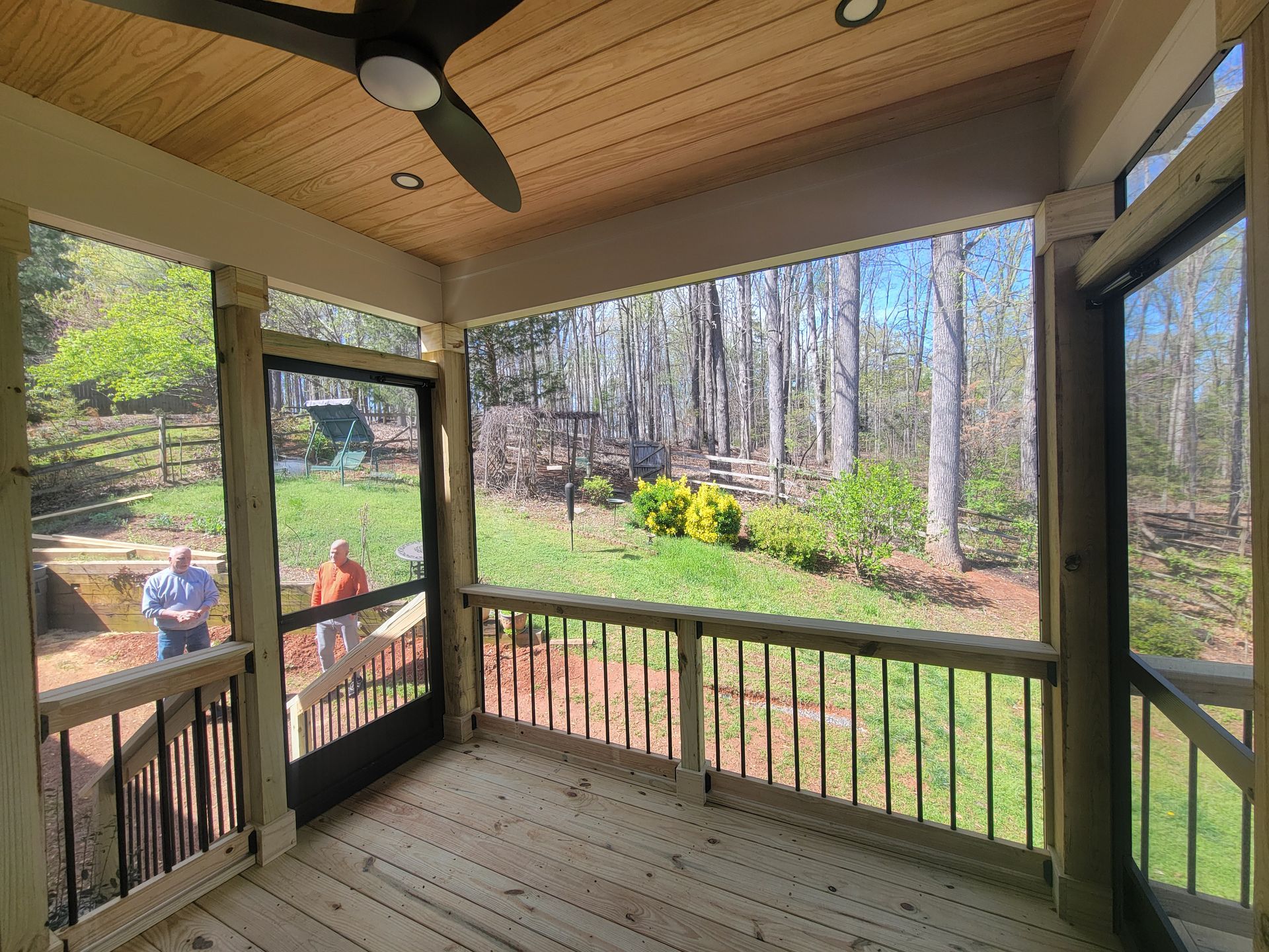 Screened porch overlooking a wooded yard and fire pit, with two people seated outside.