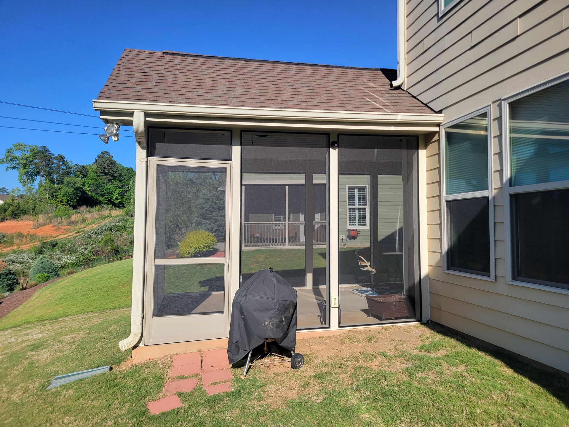 Screened patio attached to a tan house, with a covered grill and brick pavers on the grass.