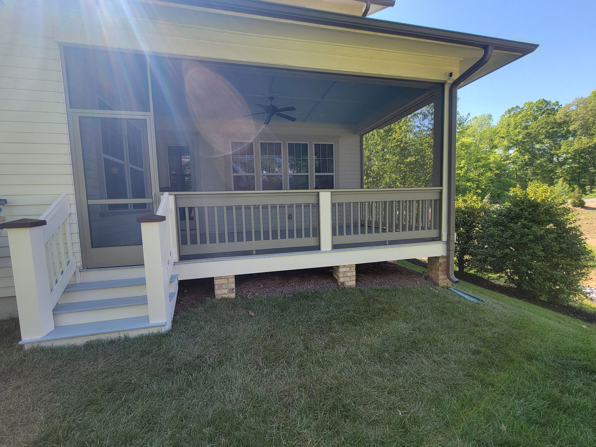 Covered front porch with white railing, gray siding, steps, and a grassy yard in sunlight