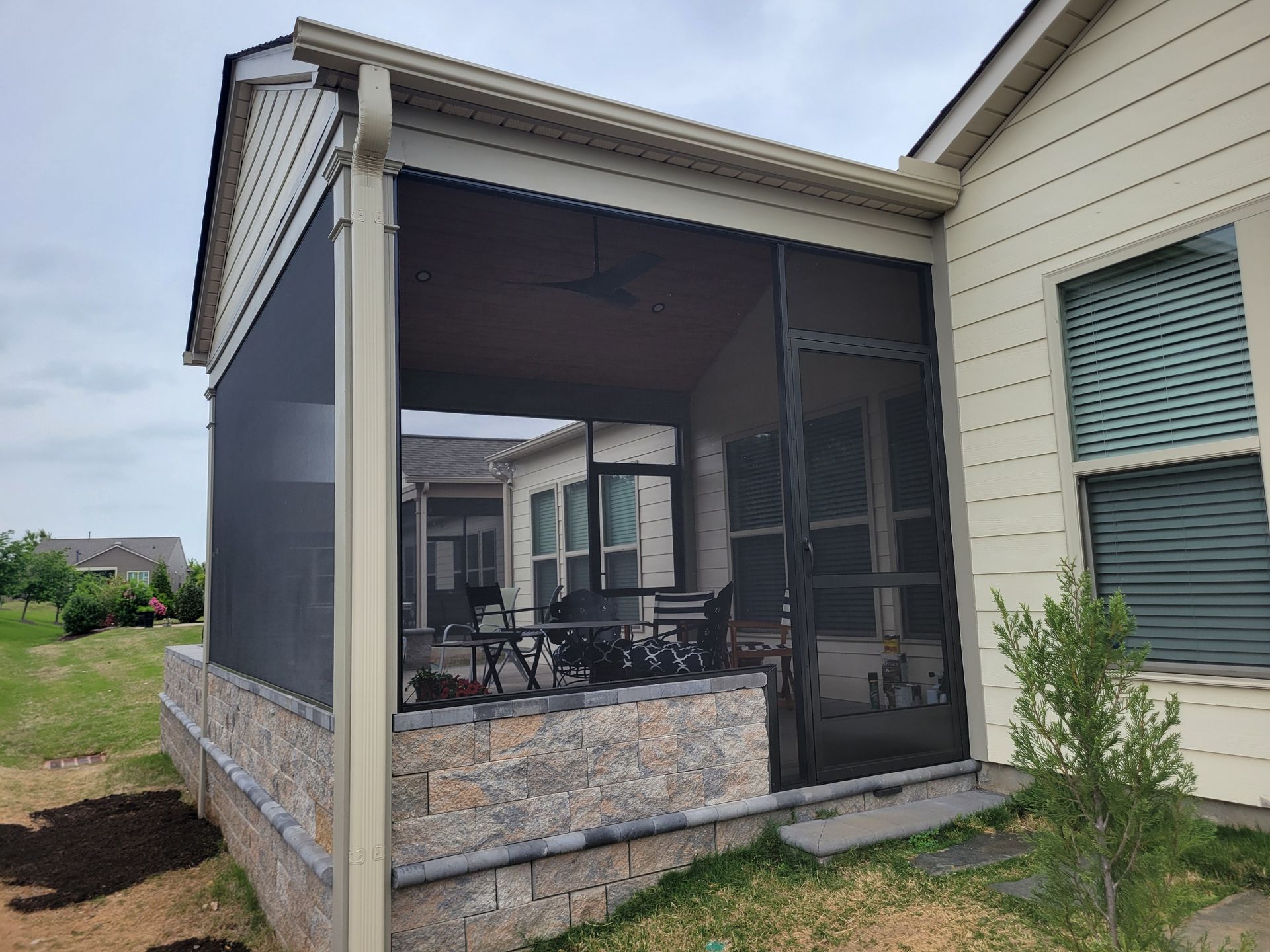 Covered patio with screened walls, outdoor furniture, and stone base beside a house.