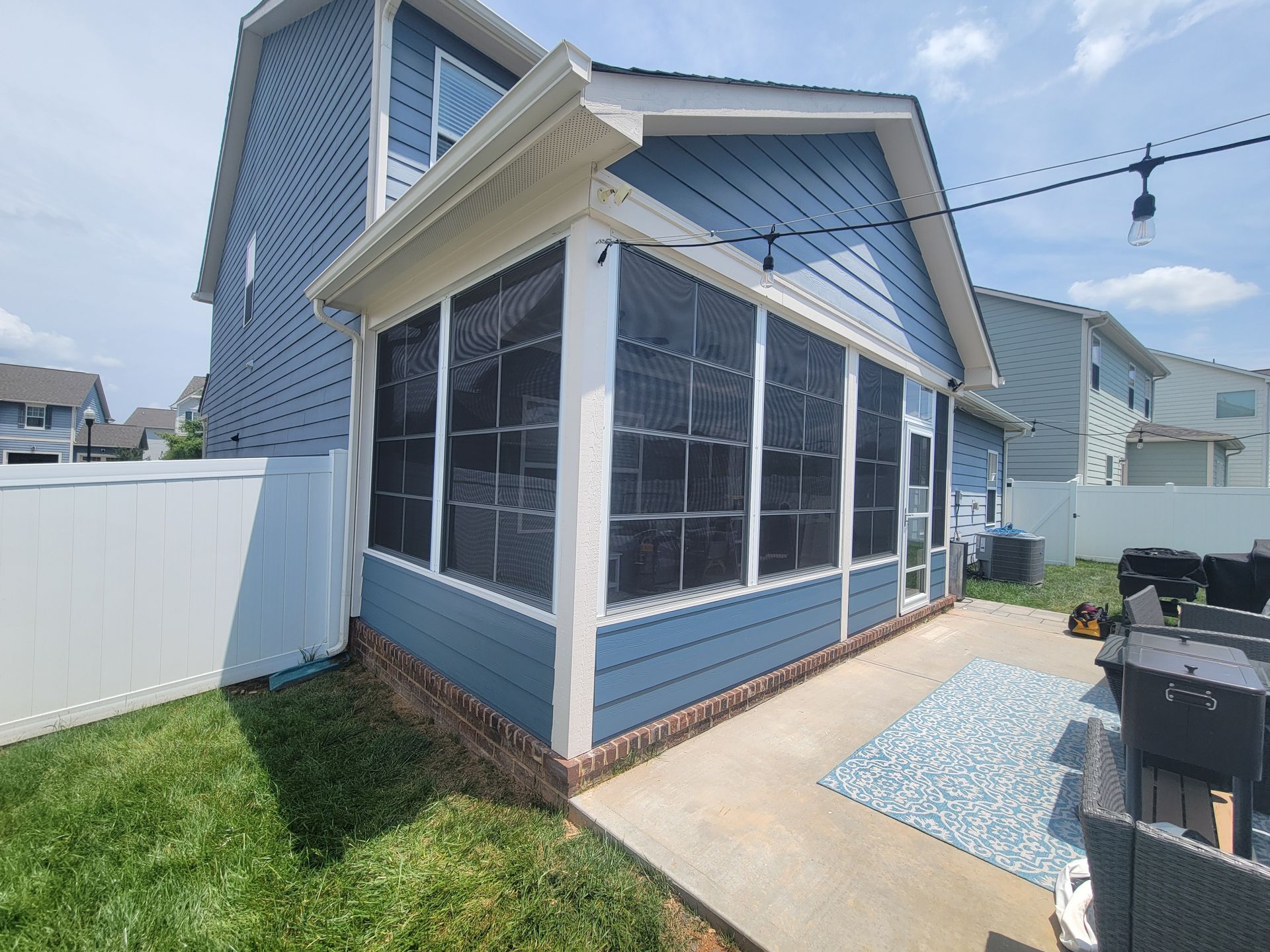 Blue screened porch attached to a house, with a white fence and small patio area beside it.