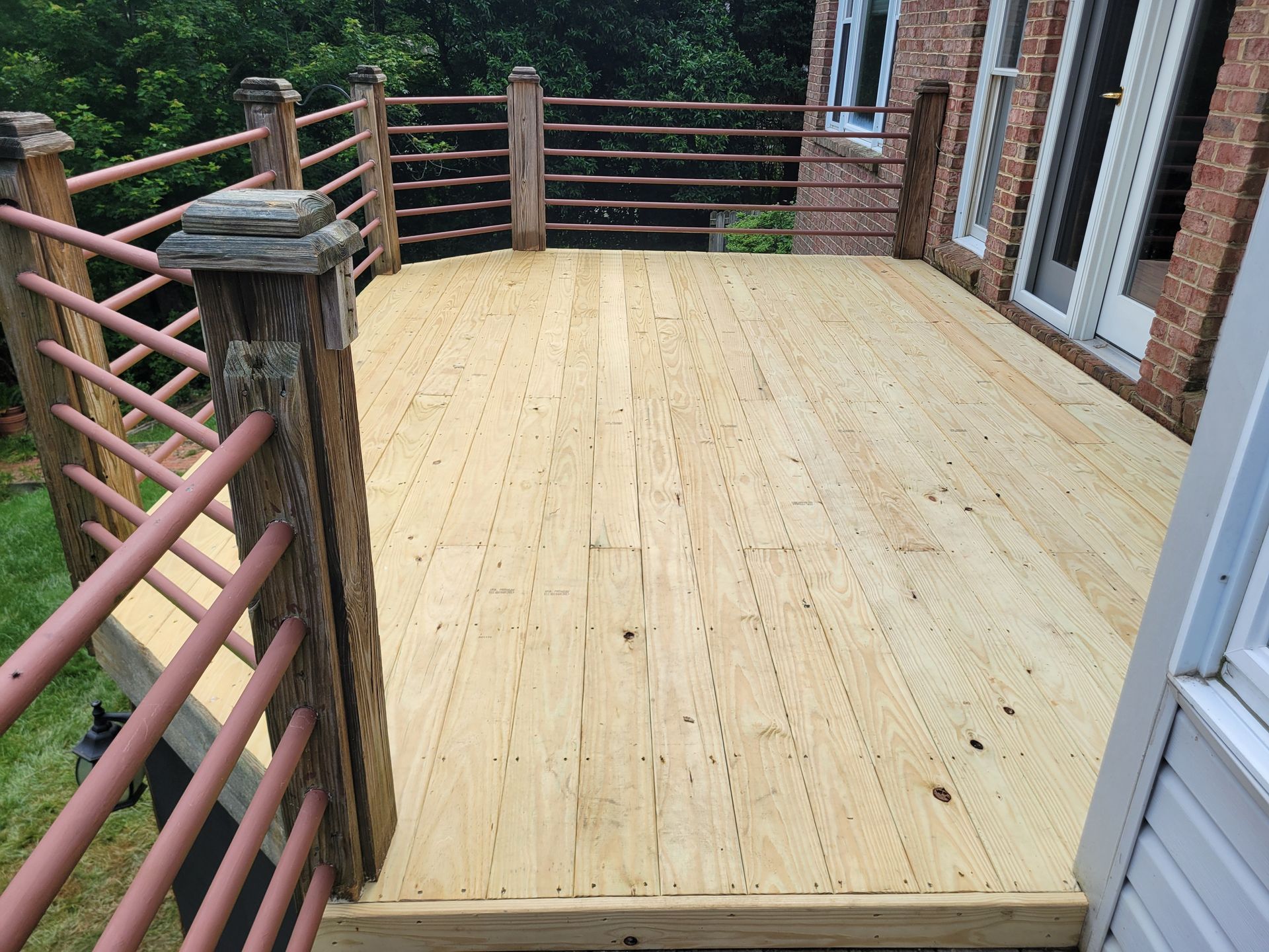Wooden deck with railings beside a brick house, overlooking trees in the background