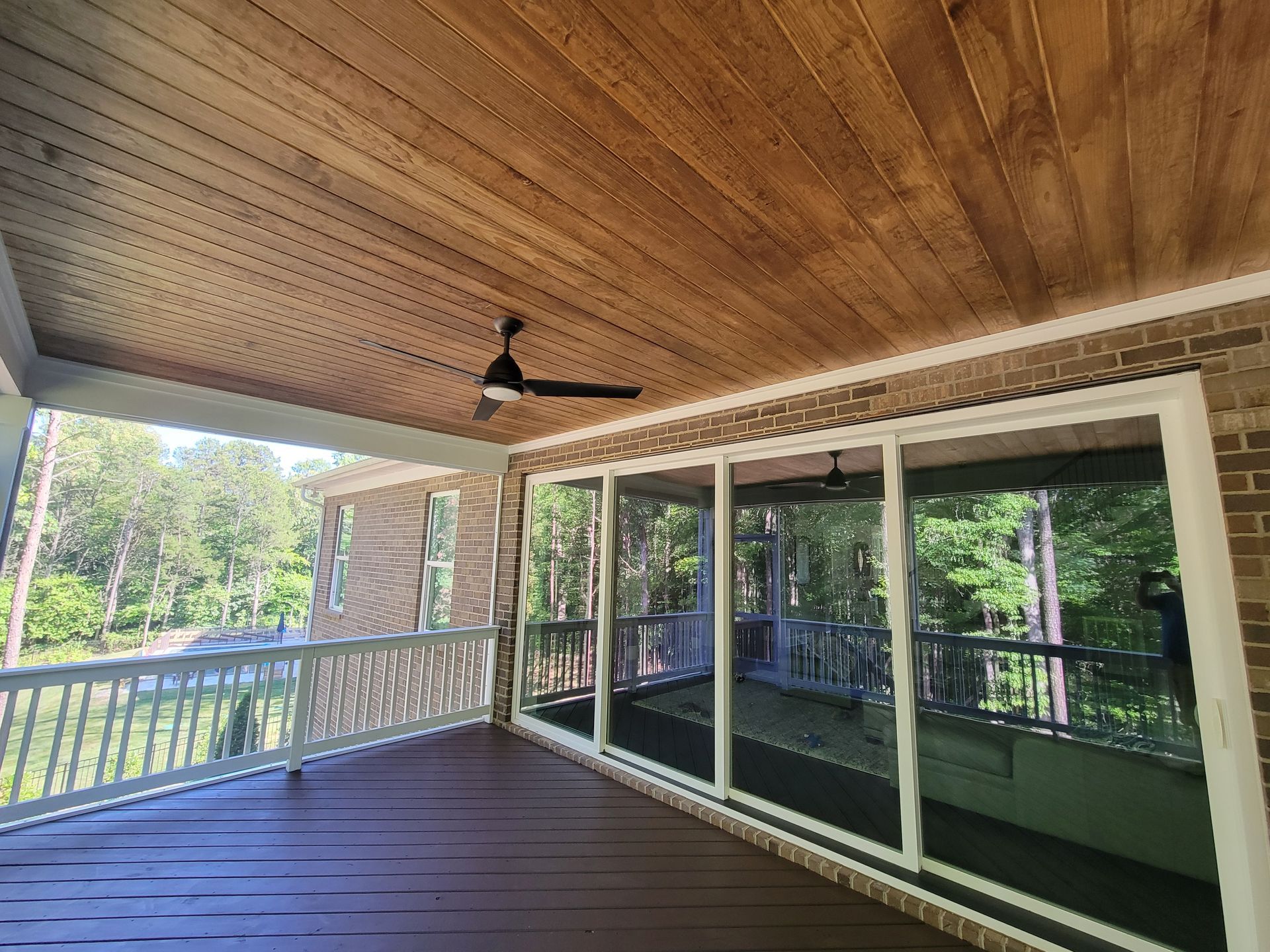 Covered wooden porch with ceiling fan, railing, and large sliding glass doors overlooking trees