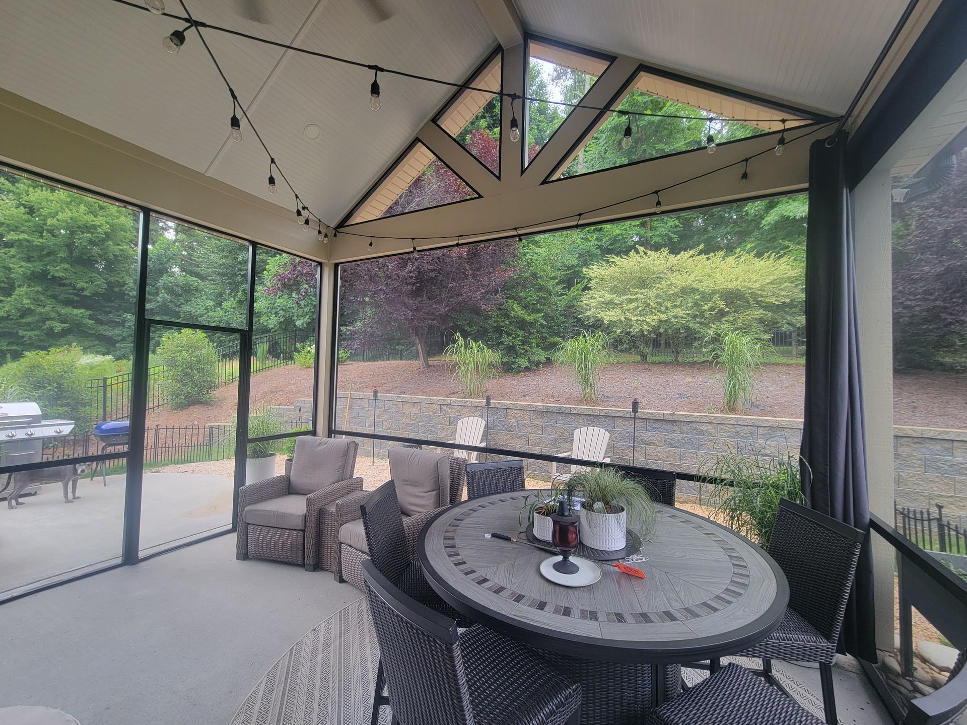 Covered patio with wicker table, cushioned chairs, and string lights overlooking a garden and hillside.