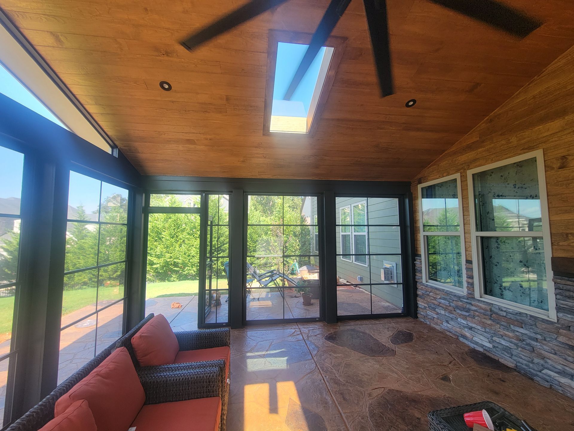 Screened porch with wood ceiling, ceiling fan, skylights, and large windows overlooking greenery.