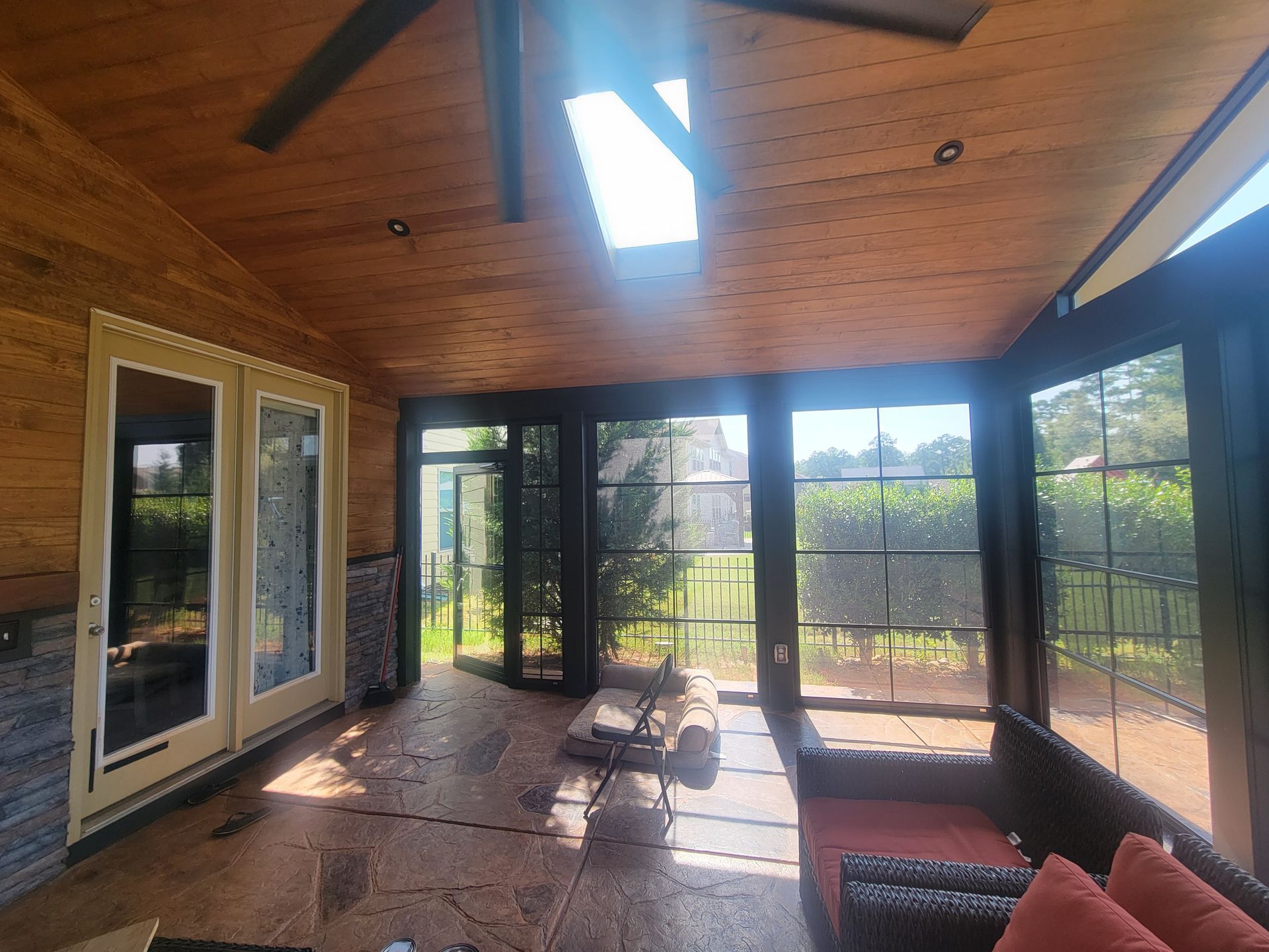 Sunlit screened porch with wood ceiling, glass doors, and wicker seating overlooking a fenced yard.