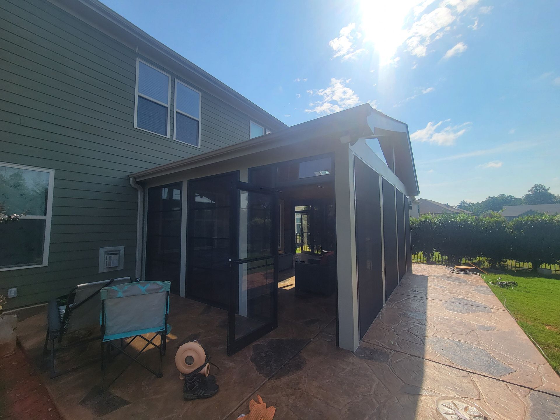 Backyard patio beside a house, with screened enclosure, chair, and garden hose in bright sunlight
