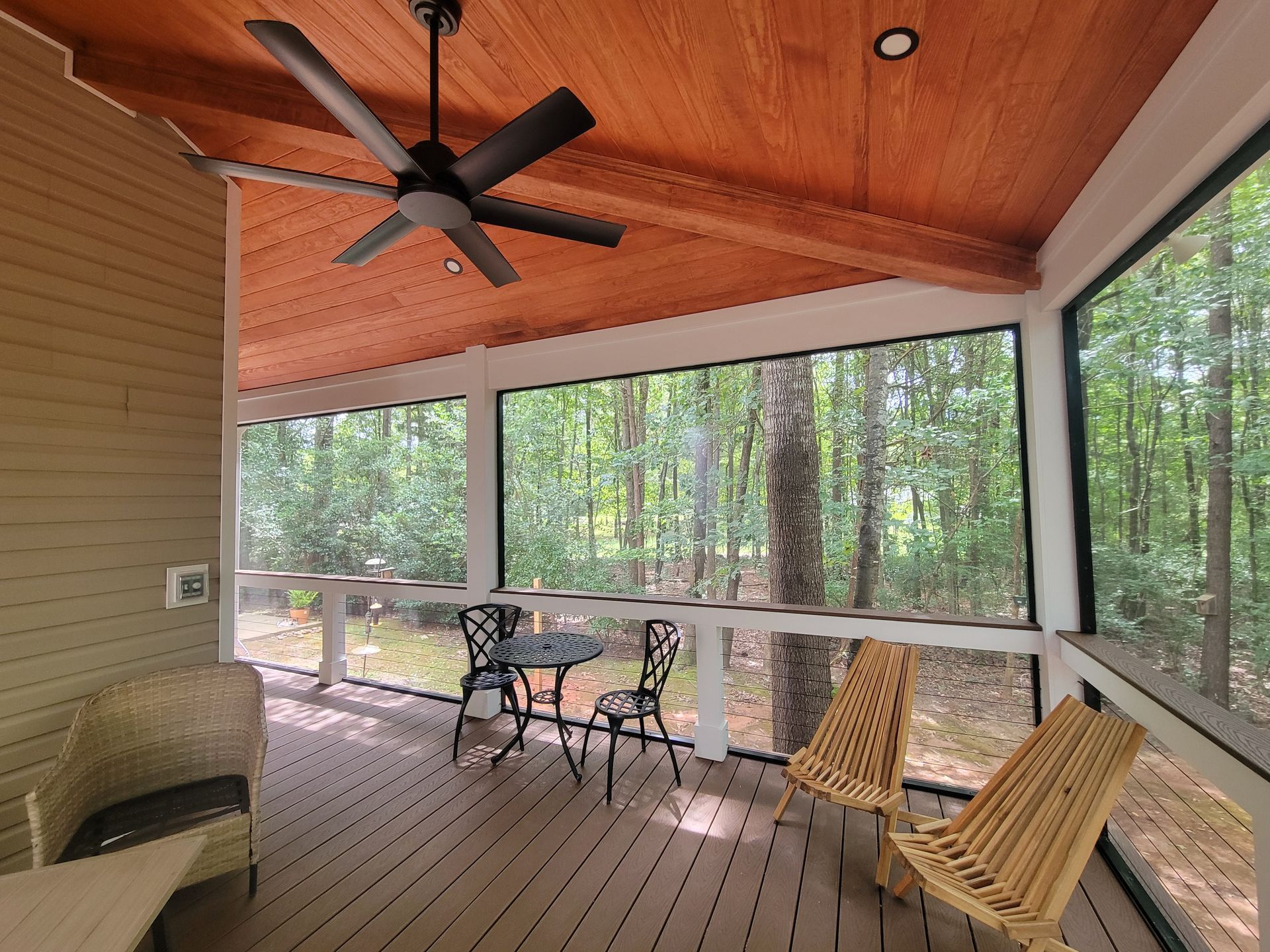 Screened porch with wood ceiling, ceiling fan, wicker chairs, and table overlooking a wooded area