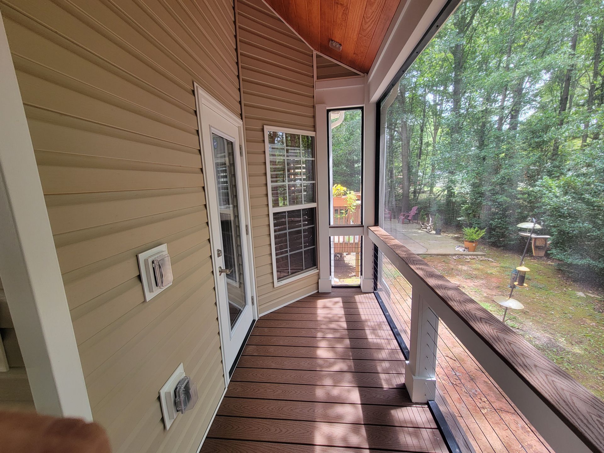 Covered wooden porch with tan siding, railing, and screened door overlooking a wooded yard.