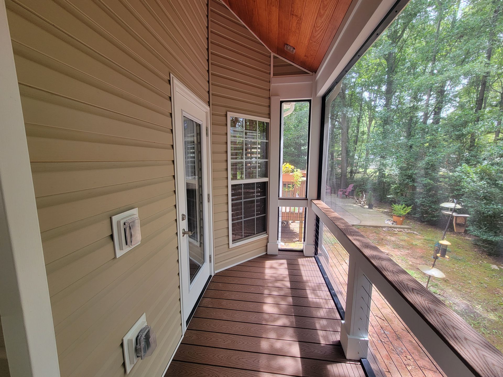 Covered wooden porch with tan siding, screened door, railing, and a wooded yard beyond.