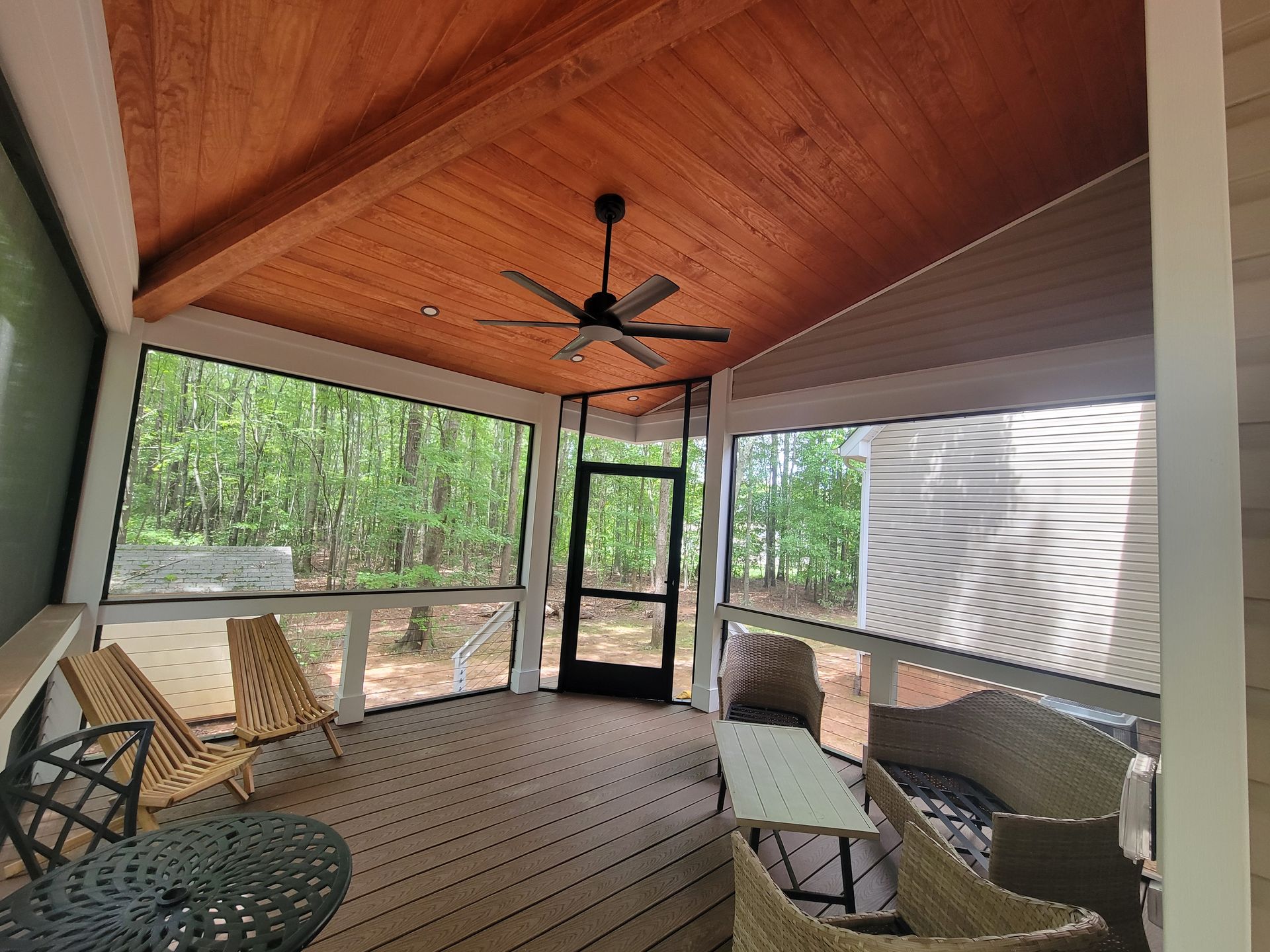 Covered screened porch with wood ceiling, ceiling fan, chairs, and a table overlooking trees.