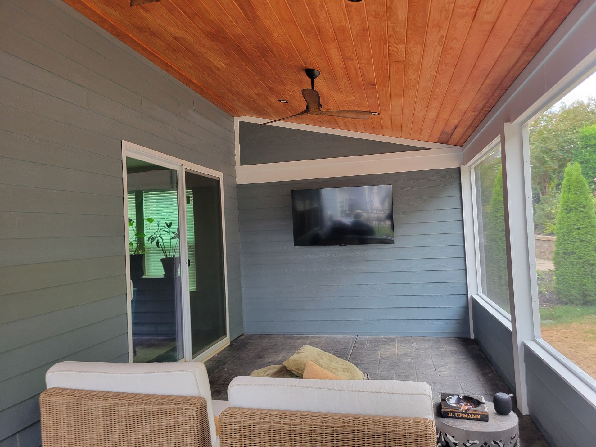 Covered patio with wood ceiling, gray walls, wall-mounted TV, and wicker seating overlooking a yard