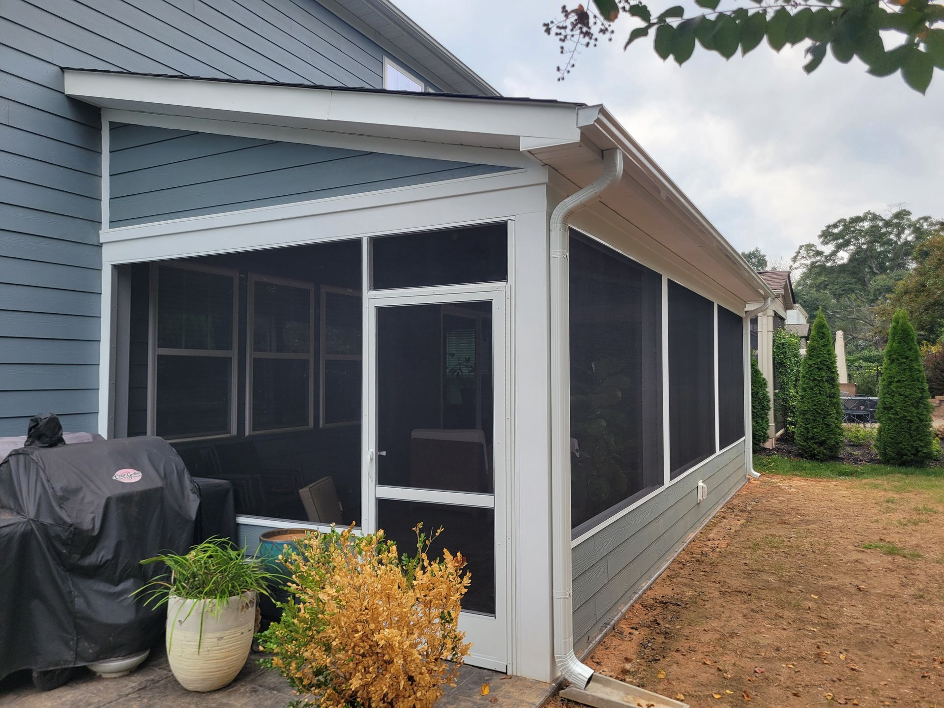 Screened porch on a gray house with a grill, potted plant, and screened windows beside a dirt yard